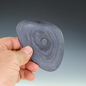 May include: A gray, organic-shaped pottery rib tool with a small, centered hole. The tool has a wood grain pattern and is held in a hand against a neutral background. This tool is used for shaping and smoothing clay.