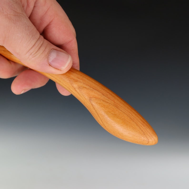 May include: A close-up of a wooden butter knife with a smooth, rounded handle. The knife is made of cherry wood and has a natural finish.