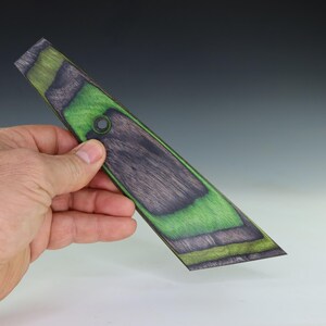 May include: A wooden, angled piece with a hole, featuring a striking color scheme. The design incorporates shades of green, black, and gray, creating a visually appealing contrast. The piece is held in a hand, showcasing its size and shape.