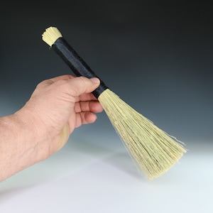 May include: A hand holds a small, natural fiber brush with a black handle. The brush is made of dried grasses and has a long, narrow shape.