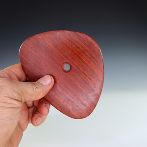 May include: A reddish-brown wooden pottery rib with a smooth, curved shape and a central hole. The rib is held in a hand, showcasing the wood grain and the tool's design for shaping clay.