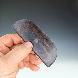 May include: A dark brown, wooden pottery rib tool with a rounded end and a small hole. The tool has a smooth, curved shape and is held in a hand against a neutral background. The wood grain is visible, adding texture to the tool.