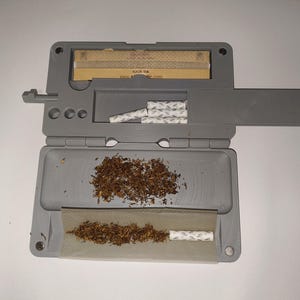 Puede incluir: Máquina de liar gris con tabaco, un cigarrillo parcialmente liado, papel de fumar y filtros. La imagen muestra el proceso de liar un cigarrillo. La máquina está abierta.
