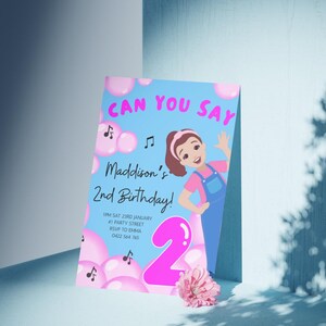 Pu&ograve; includere: Un invito di compleanno blu e rosa con il testo "Can You Say" e "Maddison's 2nd Birthday!" L'invito presenta una ragazza dei cartoni animati con i capelli castani e una camicia rosa, che tiene un palloncino rosa numero 2. L'invito include anche note musicali e bolle rosa.