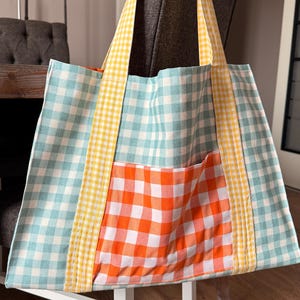 Peut inclure: Un sac fourre-tout patchwork avec un motif à carreaux vert, jaune et orange. Le sac a une grande poche sur le devant et deux anses jaunes.