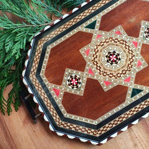 Op de afbeelding: Achtekante houten dienblad met ingelegde geometrische patronen in tinten van bruin, zwart, wit, rood en groen. Het dienblad heeft een verhoogde rand en een gladde, gepolijste afwerking.