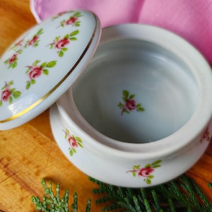 May include: A white porcelain trinket box with a lid decorated with a floral pattern of pink roses. The box has a gold trim around the edge of the lid.