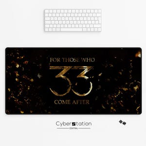 Könnte beinhalten: Schwarze rechteckige Schreibtischunterlage mit goldenem Text, der lautet: „FOR THOSE WHO 33 COME AFTER“. Die Matte hat ein goldenes Konfetti-Design und das Cyberstation Central-Logo. Über der Matte befindet sich eine weiße Tastatur.