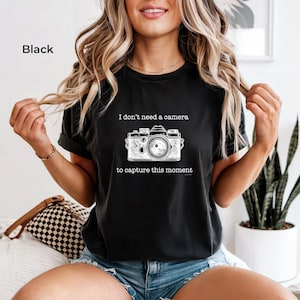May include: Black t-shirt with the text "I don't need a camera to capture this moment" and a detailed illustration of a vintage camera. The shirt is a dark color and has short sleeves.