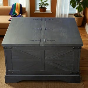 May include: A large, dark gray wooden storage chest with a hinged lid. The chest features metal hinges and corner accents, and a decorative panel design on the front. The chest is placed on a wooden floor.