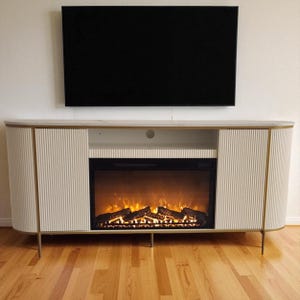 May include: A modern off-white entertainment center with a built-in electric fireplace and a black television mounted above. The cabinet has a ribbed design with gold accents and legs. The fireplace displays a realistic flame effect.