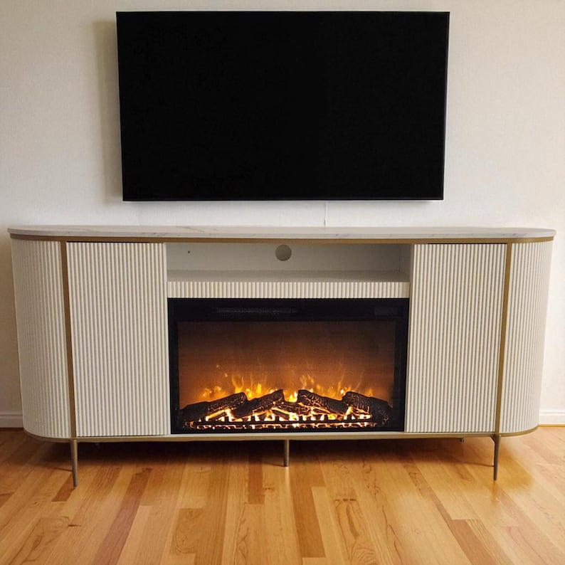 May include: A modern off-white entertainment center with a built-in electric fireplace and a black television mounted above. The cabinet has a ribbed design with gold accents and legs. The fireplace displays a realistic flame effect.