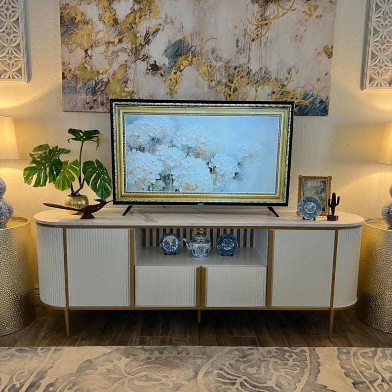 May include: A modern white and gold TV stand with a marble top, gold trim, and ribbed cabinet doors. The stand holds a flat-screen TV with a gold frame, decorative items, and a houseplant. The room features a large abstract painting.