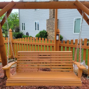 May include: A wooden swing set with a brown finish, featuring a slatted seat and backrest. The swing hangs from chains attached to a wooden frame. The background includes a wooden fence and a house.