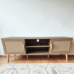 May include: A light brown wooden TV stand with two doors and an open center section. The doors have woven cane panels. The stand has a mid-century modern design with angled legs and wooden handles. The stand is approximately 60 inches long.