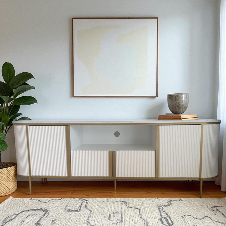 May include: A modern white and gold media console with fluted door panels and a marble top. The console has gold legs and trim, and a central open shelf. A framed abstract artwork hangs above the console.