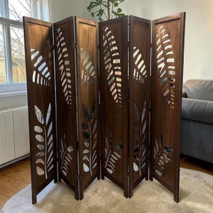 May include: A dark brown, six-panel room divider with leaf-shaped cutouts. The wooden screen stands on a white rug, with a window and a couch visible in the background. The leaves are cut out of the wood, creating a decorative design.