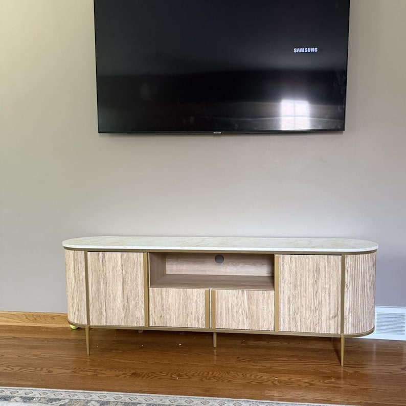 May include: A light wood-grain TV stand with a marble top and gold-colored metal legs. The stand has two sets of doors and a central open shelf. A large black Samsung television is mounted on the wall above the stand.