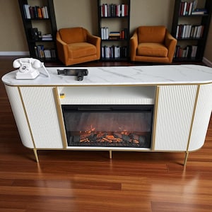 May include: A white and gold entertainment center with a marble top, vertical ribbed sides, and a built-in electric fireplace. The center has a gold trim and legs. A white rotary phone and a black object are on the top.