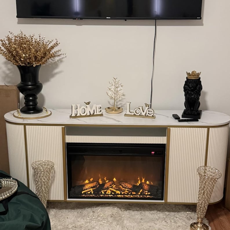 May include: A white and gold entertainment center with a marble top, featuring a built-in electric fireplace. Decorative items include a black vase with gold accents, a lion statue, and the words "Home" and "Love".