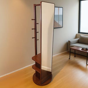 May include: A dark brown wooden standing mirror with a full-length, off-white mirror. The frame has a coat rack with pegs and two shelves. The mirror is on a round base. The design is modern and minimalist.