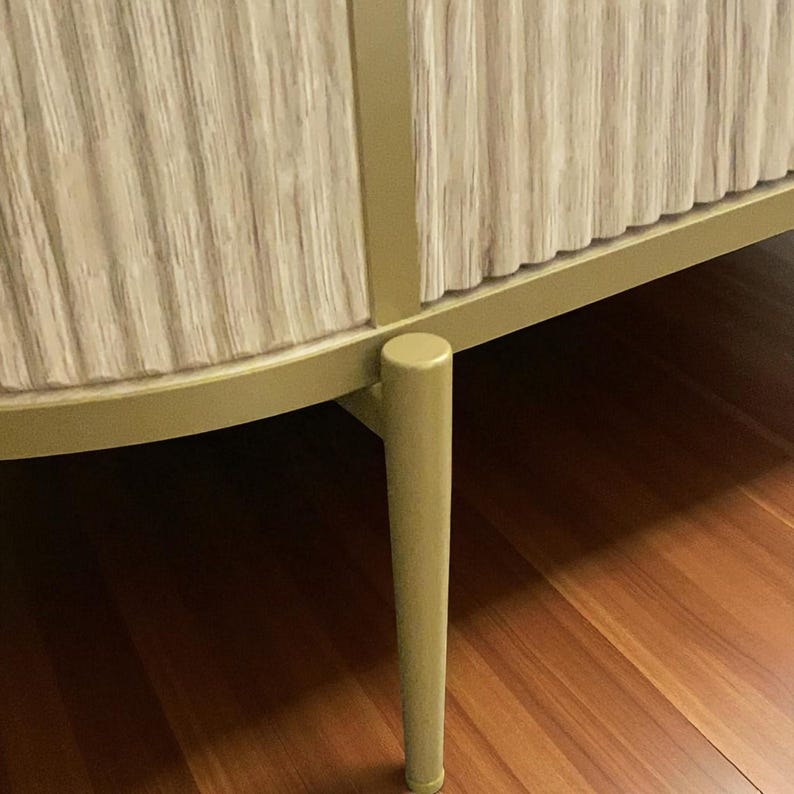 May include: A close-up of a cabinet with a light beige, vertically ribbed front. The cabinet has a gold-colored frame and legs. The legs are cylindrical and slightly tapered. The cabinet is set on a wooden floor.