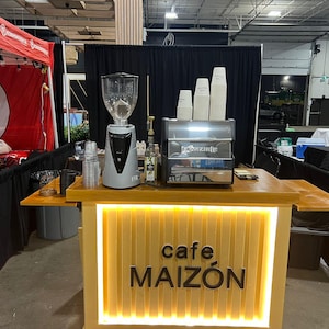 May include: A coffee bar with a light wood counter and a sign that reads "cafe MAIZÓN". The bar includes a coffee grinder, espresso machine, and stacks of paper cups. The bar is set up at an event.