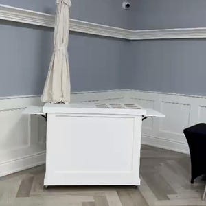 May include: A white bar with a beige parasol is set up in a room with grey walls and a light wood floor. The bar has a white countertop and a white cabinet. The parasol is closed and positioned above the bar.