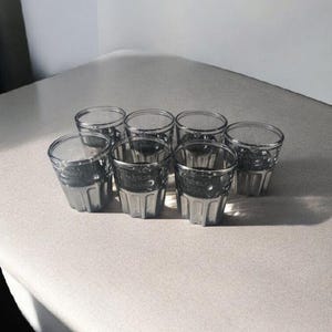 Puede incluir: Conjunto de ocho vasos de chupito de vidrio transparente con un acabado texturizado y esmerilado. Los vasos están dispuestos en dos filas de cuatro sobre una superficie blanca.