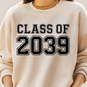Puede incluir: Una sudadera color crema con el texto "CLASS OF 2039" en letras negras en negrita. La sudadera tiene cuello redondo y mangas largas. El texto está delineado en blanco.