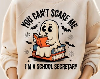 May include: Beige sweatshirt with a Halloween-themed graphic. The design features a cartoon ghost reading a book, with the text "YOU CAN'T SCARE ME I'M A SCHOOL SECRETARY" above and below the ghost. Bats and spiderwebs complete the design.
