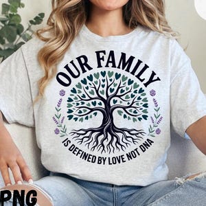 May include: Light gray t-shirt with a family tree design. The tree has heart-shaped leaves in shades of green and blue. The text "OUR FAMILY IS DEFINED BY LOVE NOT DNA" is printed in black.