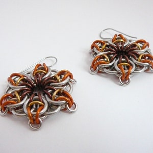 May include: A pair of handmade earrings featuring a floral design. The earrings are crafted from interwoven metal rings in silver, copper, bronze, and gold tones. Each earring has a silver hook for pierced ears.
