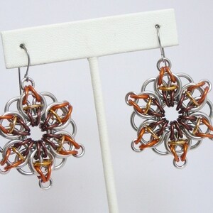 May include: A pair of handmade earrings featuring a floral design. The earrings are constructed from silver-colored metal rings, with accents of copper, bronze, and gold-colored rings. The earrings are displayed on a white earring stand.