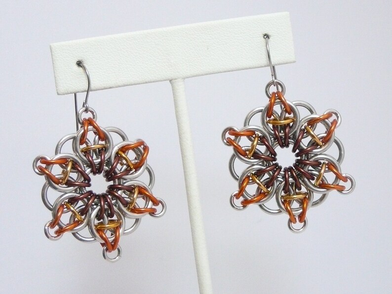 May include: A pair of handmade earrings featuring a floral design. The earrings are constructed from silver-colored metal rings, with accents of copper, bronze, and gold-colored rings. The earrings are displayed on a white earring stand.
