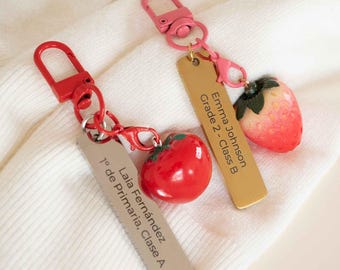 May include: Two keychains with red and pink accents. One keychain has a red tomato charm and a silver tag with the text "Laia Fernández 1º de Primaria, Clase A". The other keychain has a strawberry charm and a gold tag with the text "Emma Johnson Grade 2 - Class B".