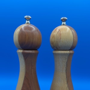 May include: Two wooden salt and pepper mills featuring a curved, hourglass-shaped body with alternating light and dark wood grain segments. Each mill is topped with a small, polished silver-colored metal knob for adjusting the grinding mechanism.