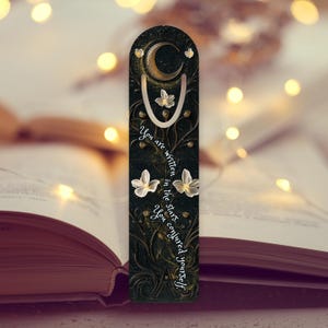 May include: A dark green bookmark with a gold crescent moon and white floral accents. The bookmark has the text "You are written in the stars. You conjured yourself."
