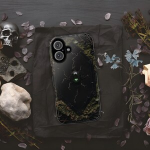 Peut inclure: Coque de smartphone noire avec un design gothique. Le boîtier présente un panneau central noir avec une gemme verte et des détails complexes. Le boîtier a un fond sombre et texturé avec un crâne et des éléments floraux.