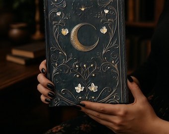 May include: A dark teal journal with a gold crescent moon and floral design. The cover features embossed gold embellishments and white flowers. The journal is held in a person's hands.