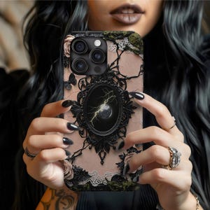 Peut inclure: Coque de téléphone décorative avec un cadre noir orné d'un motif de lune et d'éclairs. L'étui a un fond rose avec des roses, des papillons et des accents de dentelle. L'étui est conçu pour un smartphone.