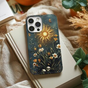 May include: A phone case with a dark blue background featuring a yellow sun, white flowers, and two white skulls. The case has a camera lens cutout and a dark blue border.