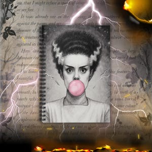 May include: A spiral-bound notebook featuring a pencil sketch of a woman with Frankenstein-like features blowing a pink bubble. The background includes text and lightning bolts, creating a gothic theme.
