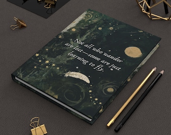Op de afbeelding: Een zwart en gouden notitieboek met een citaat dat luidt "Not all who wander are lost - some are just learning to fly." Het notitieboek heeft een marmermotief met gouden sterren en een witte veer.