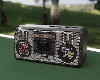 Może przedstawiać: Czarny i srebrny boombox z ozdobnymi naszywkami. Naszywki zawierają serce z napisem "I ❤️ 90s" i kwiat z symbolem pokoju. Boombox ma siatkową osłonę głośnika i retro design.