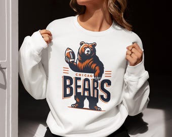 Może przedstawiać: Biała bluza z nadrukiem brązowego niedźwiedzia trzymającego piłkę do futbolu. Pod niedźwiedziem znajduje się napis "Chicago Bears".