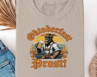 May include: Beige t-shirt featuring a cartoon gorilla in lederhosen holding a beer stein. The design includes the words "Oktoberfest" and "Prost!" in a vintage style, with a sunset backdrop.