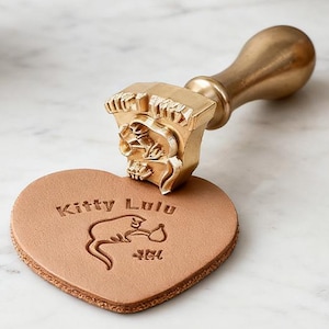 May include: A brass stamp with a wooden handle is pressed onto a heart-shaped piece of tan leather. The stamp has a detailed design, and the leather is embossed with the words "Kitty Lulu" and a cat illustration. Several small, unglazed pottery pots are in the background.