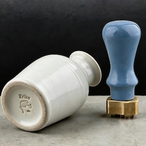 May include: A white ceramic vase with a flared rim and a blue wax seal stamp. The vase is inscribed with the name "Erika" and a small illustration. The stamp has a gold base.
