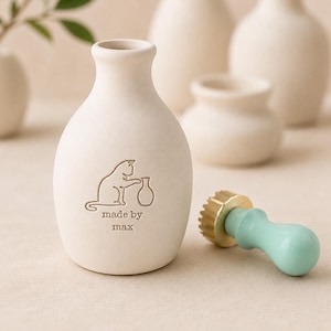 Può includere: Una bottiglia in ceramica bianco sporco opaco con un'incisione che raffigura un gatto che cerca di raggiungere un piccolo vaso, con il testo "made by max" sotto. Accanto alla bottiglia si trova un sigillo in ceralacca con manico verde menta.